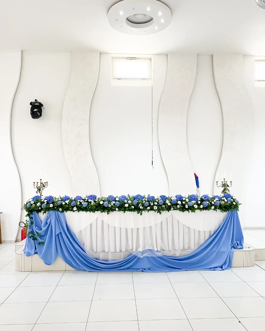 Dekoracija mladenačkog stola sa plavim i belim cvećem, svećnjacima i plavim platnom